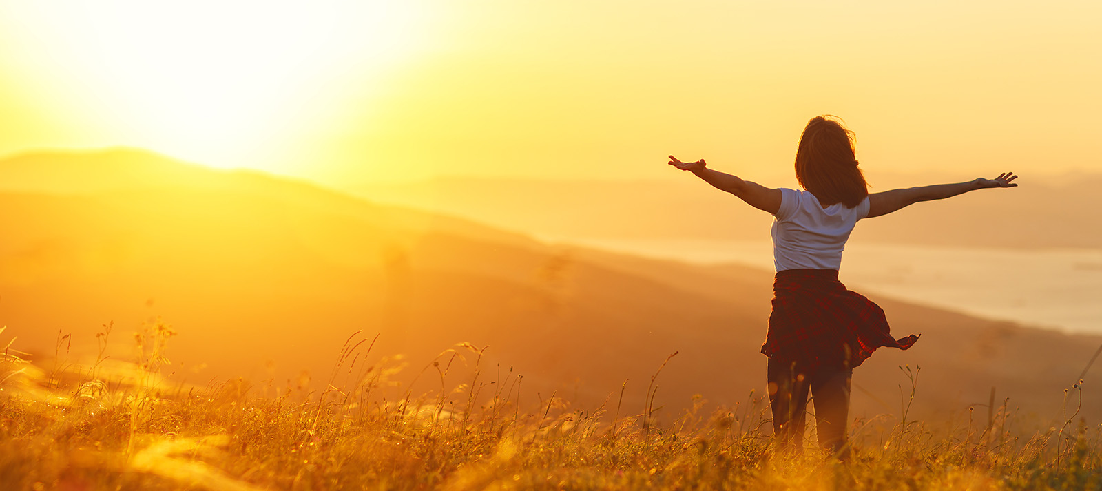 A person standing on a hilltop at sunrise with their arms raised, overlooking a valley under a clear sky.