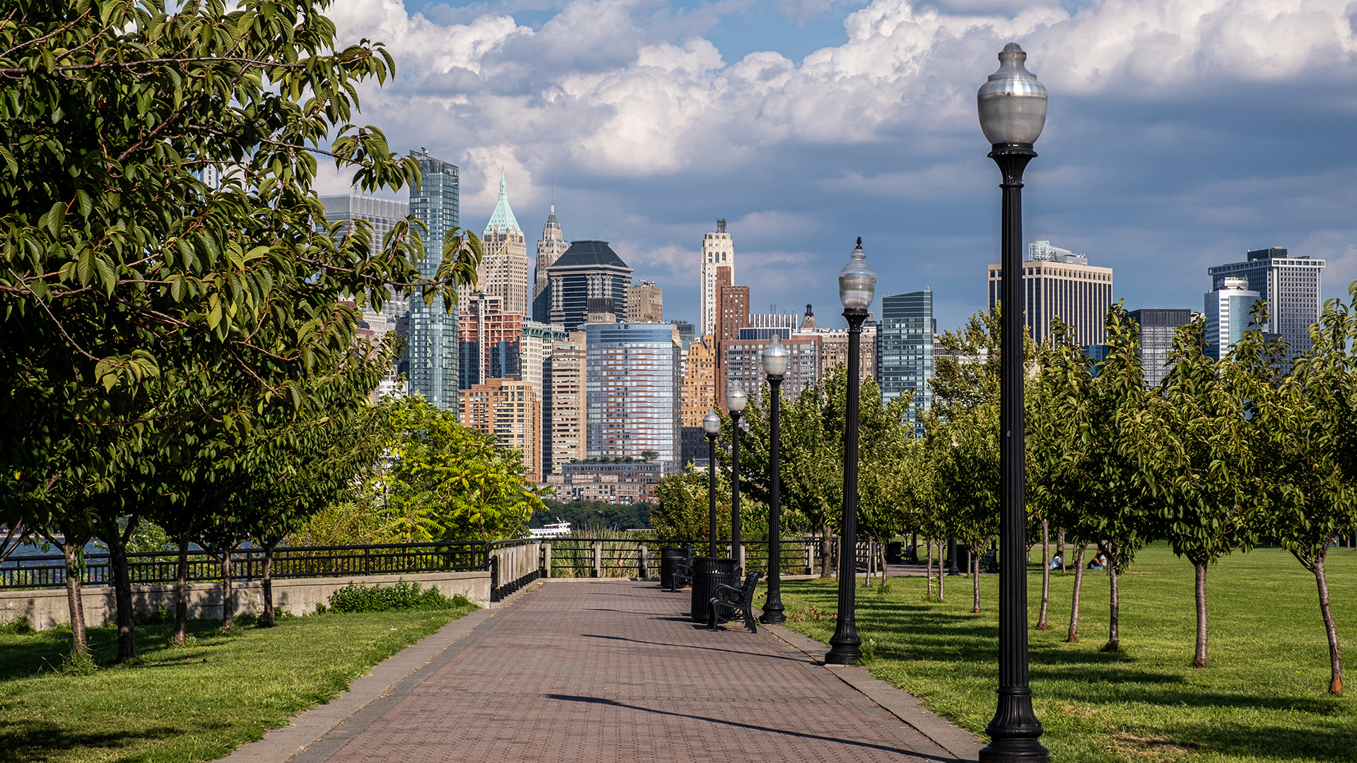 The image depicts a city skyline with a riverfront park area featuring a walkway, trees, street lamps, and a bridge leading to a downtown area with high-rise buildings under a partly cloudy sky.
