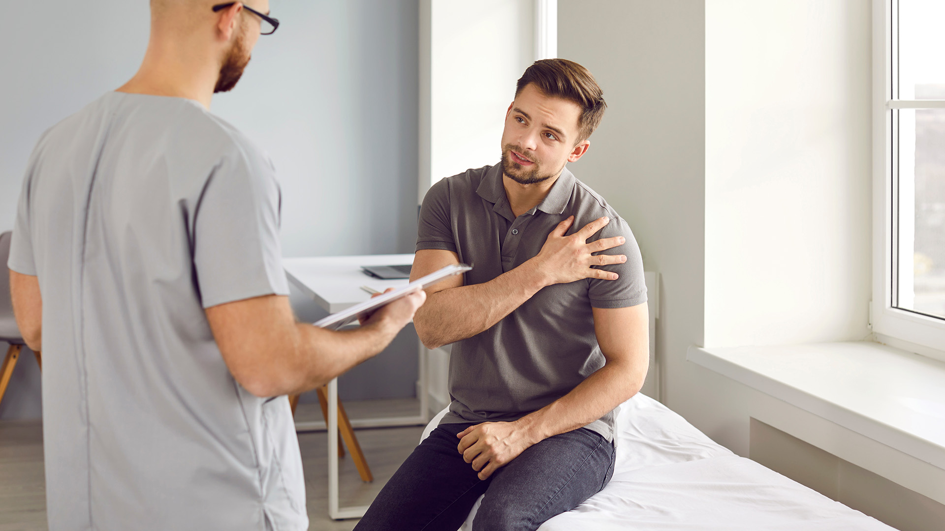 A man sitting on a bed with his arms crossed while receiving attention from a standing healthcare professional who appears to be taking notes.