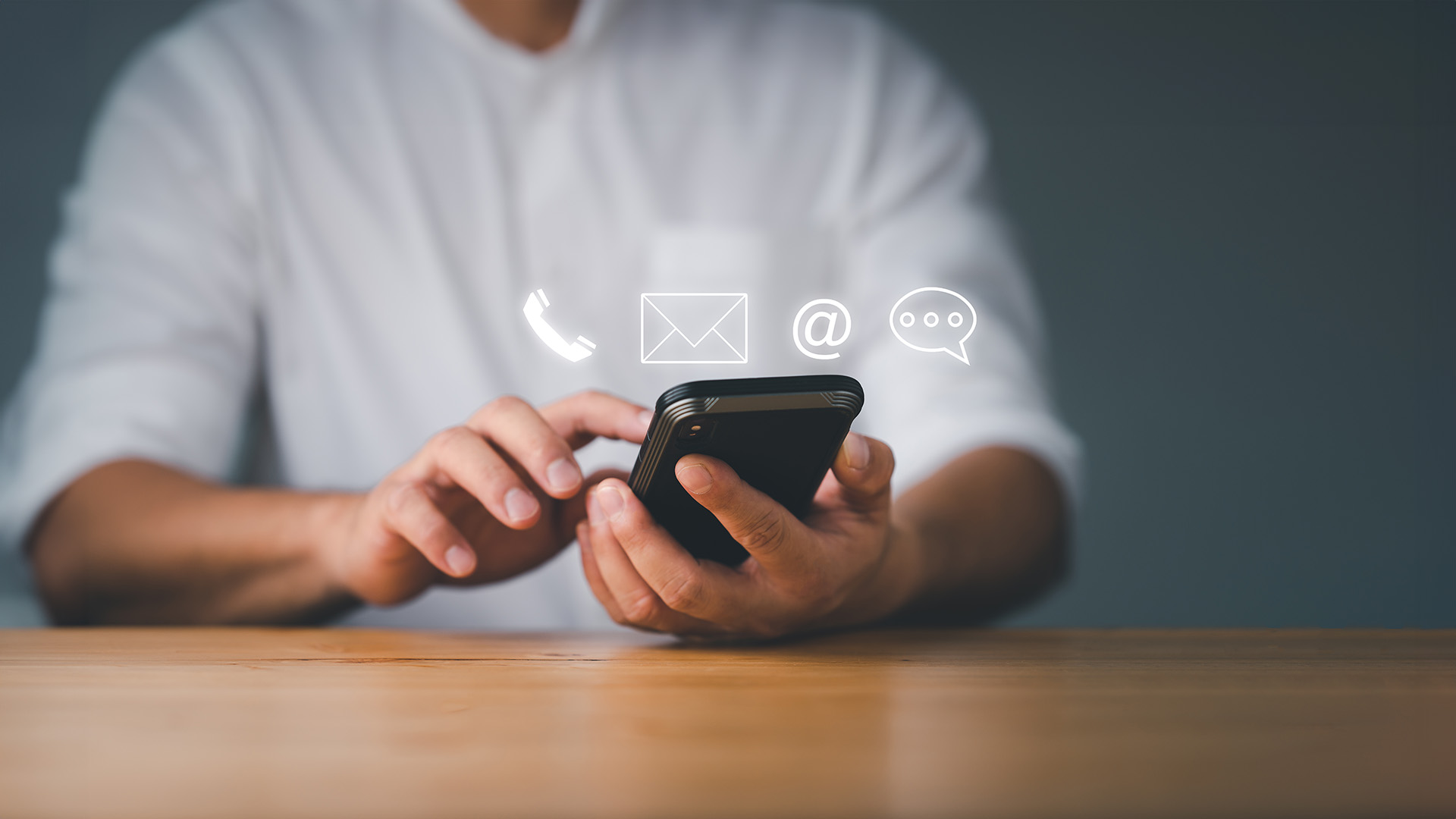 The image shows a person using a smartphone with various icons displayed on the screen, including a social media app, an email icon, and a messaging app, while sitting at a table.