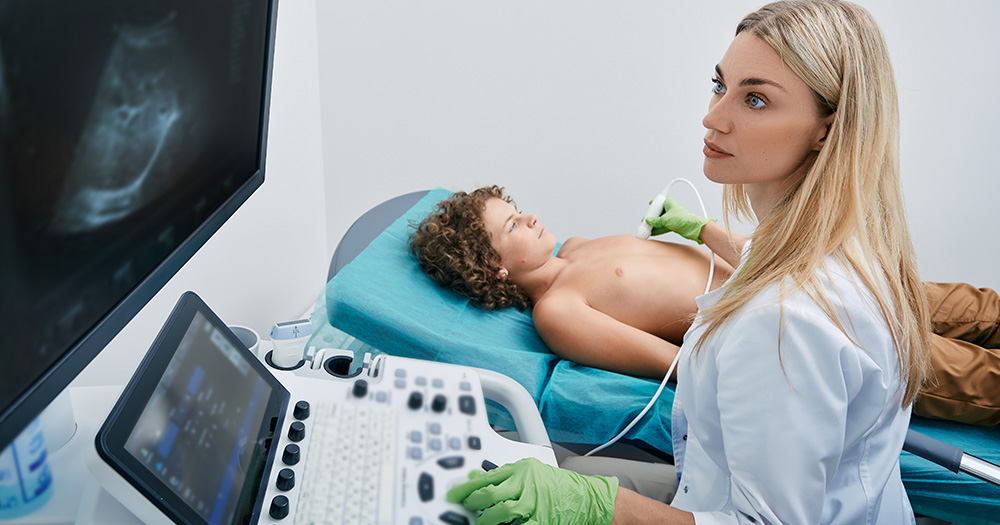 A woman wearing a white lab coat sits at a table with medical equipment, examining images on a computer screen while a young boy lies on a bed with his head resting on a pillow, receiving medical attention from her.