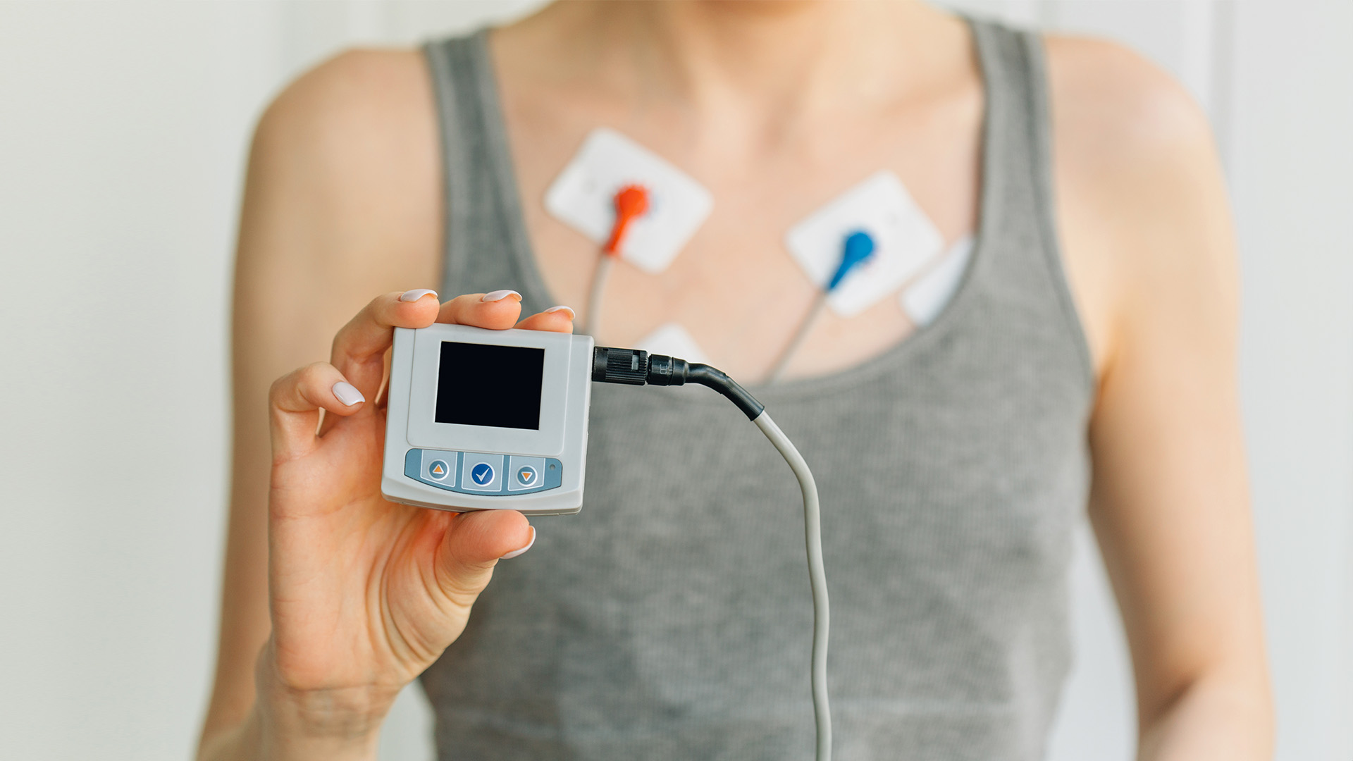 A person holding a medical device with an ECG monitor attached to their chest, displaying heart rate readings on a screen, against a white background.