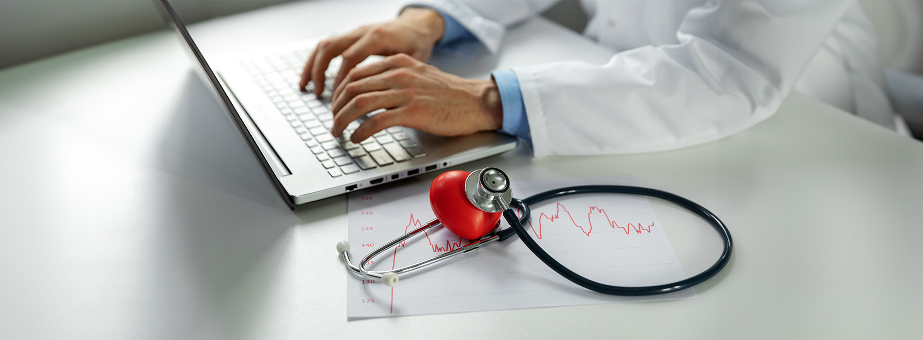 Medical professional using laptop with stethoscope and medical equipment in background.