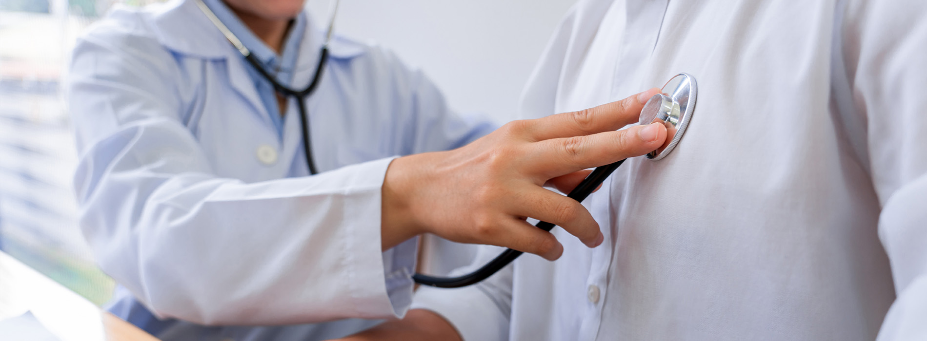 The image depicts a doctor in a white coat assisting a patient by using a stethoscope on their chest, with both individuals dressed professionally and standing indoors.