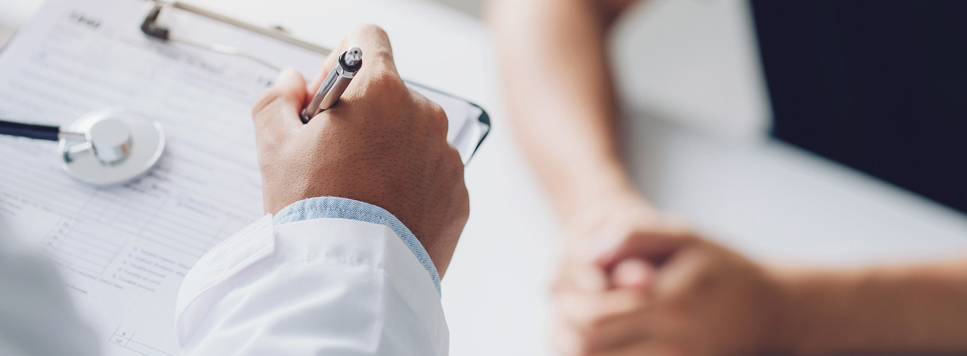 The image shows a medical professional sitting at a desk with papers, wearing a stethoscope around their neck, with a patient s hand resting on their shoulder, both hands visible, in an office setting.