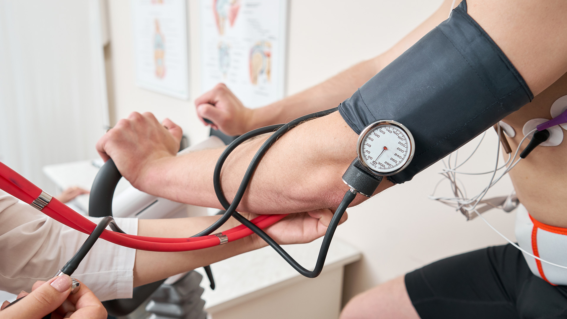 A person in a medical setting holding a blood pressure cuff and stethoscope while another individual is being monitored with a blood pressure machine.