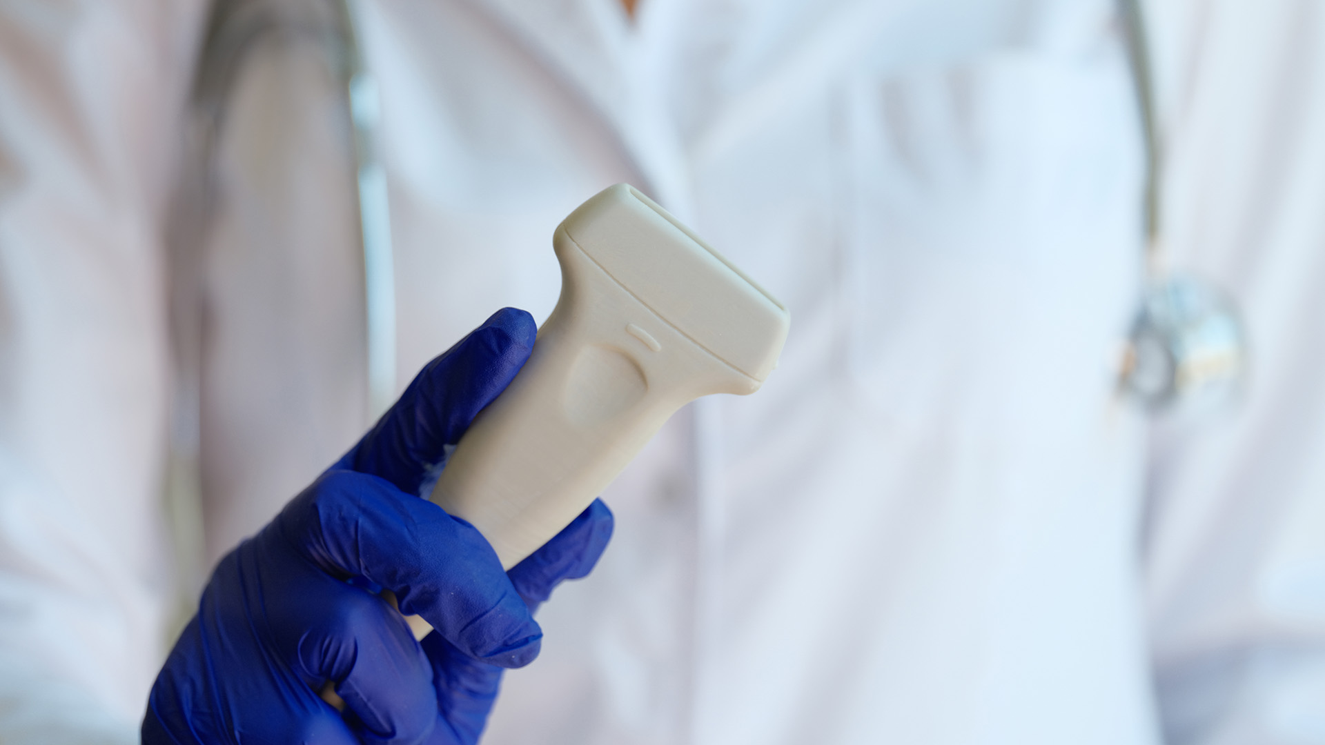 The image shows a medical professional holding a syringe with a clear barrel against a blurred background, wearing blue gloves and a stethoscope.