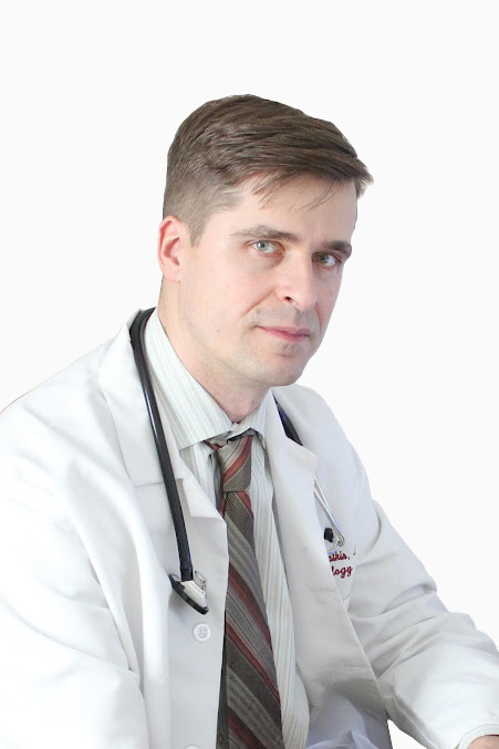 A man wearing a white coat, stethoscope, and tie, with short brown hair, posing for a headshot against a plain background.