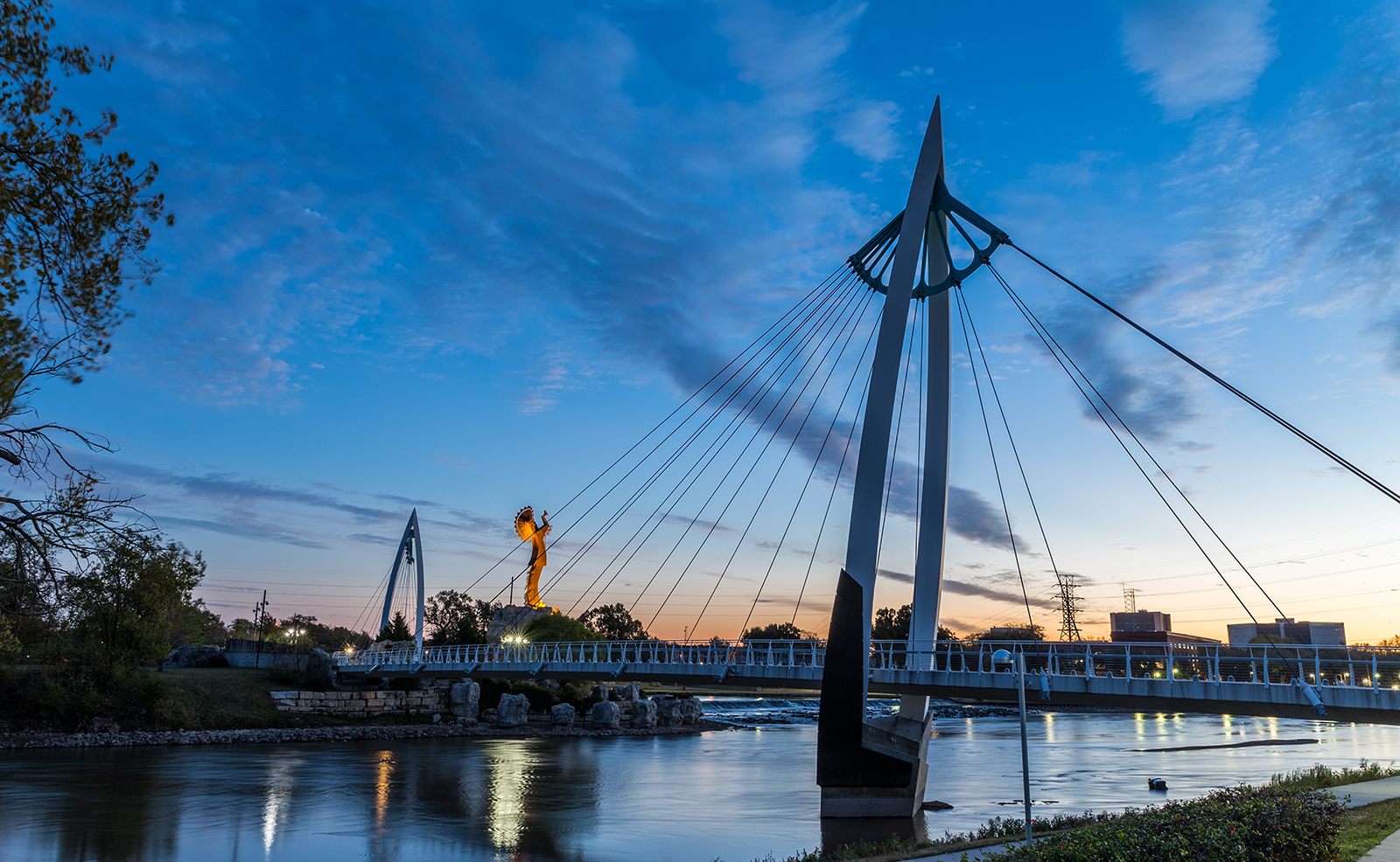 The image shows a serene nighttime scene with a bridge illuminated against a dark sky, featuring a suspension bridge with two towers connected by cables supporting a walkway, set over a calm river under a starry sky, with a cityscape in the distance and a single lamppost visible on the left.