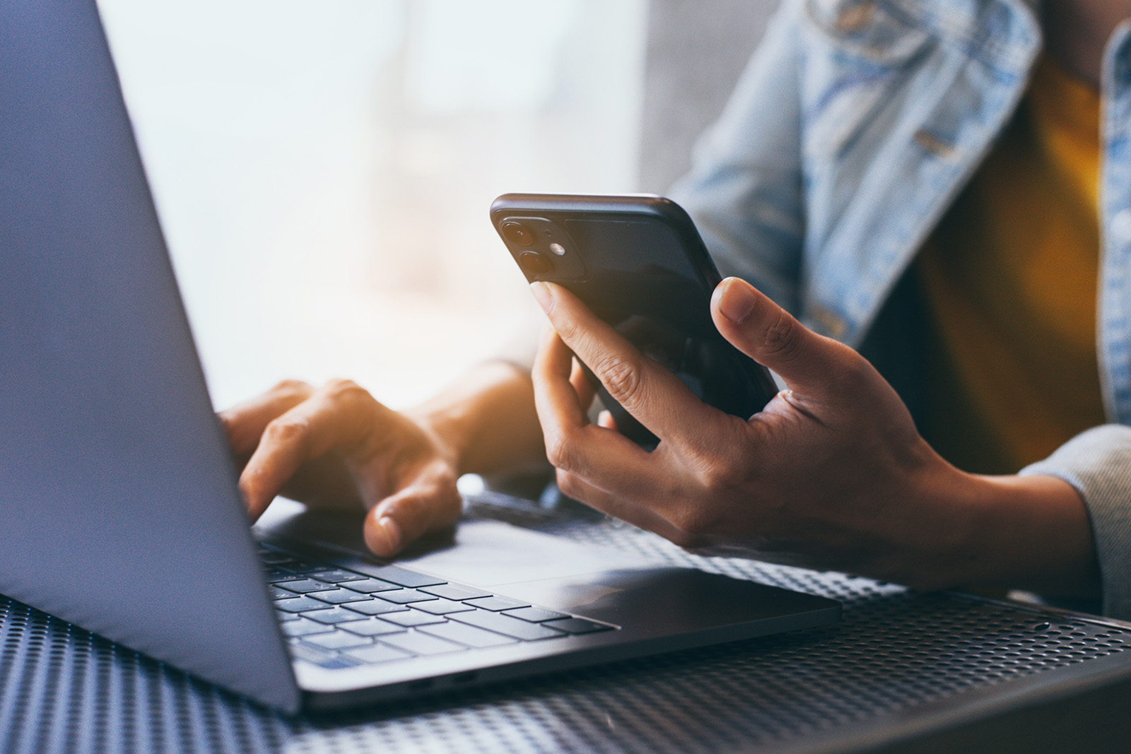 The image shows a person using a smartphone while sitting at a table with a laptop computer.