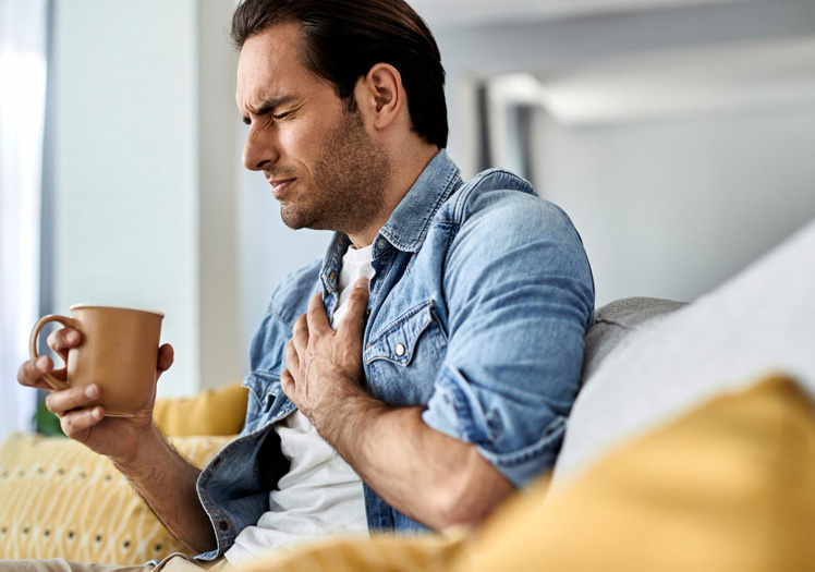 A man sitting on a couch with his hand on his chest, wearing a denim jacket and holding a coffee cup, looking concerned while possibly experiencing chest pain.