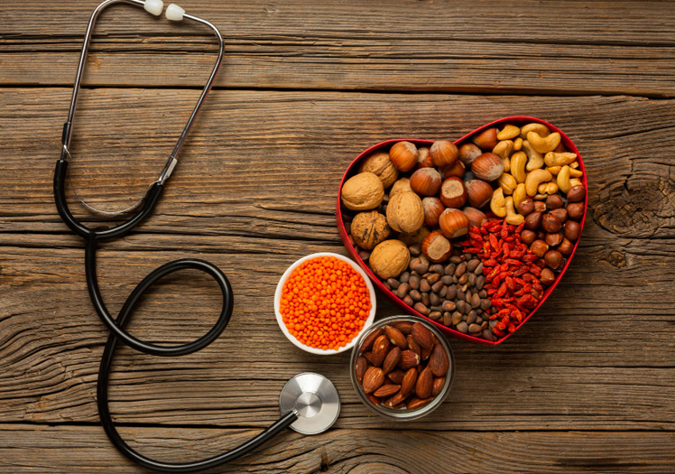 The image shows a collection of heart-shaped objects on a table, including a stethoscope, medical supplies, and various foods such as nuts and seeds, with a red heart symbolizing love or care in the center, suggesting a theme related to health or wellness.