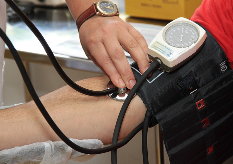 The image shows a person taking a blood pressure reading using a cuff on an arm with a visible vein, while another individual is seated nearby with their leg elevated and wearing a compression stocking.