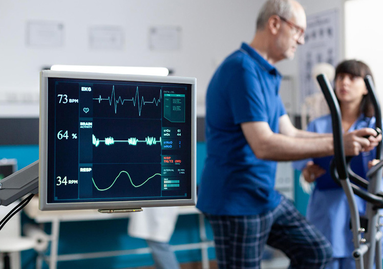 In the image, there is an elderly man using a stationary bicycle with an attached monitor displaying various health data, such as heart rate and blood pressure, while a woman, presumably a healthcare professional, observes him in a medical setting.