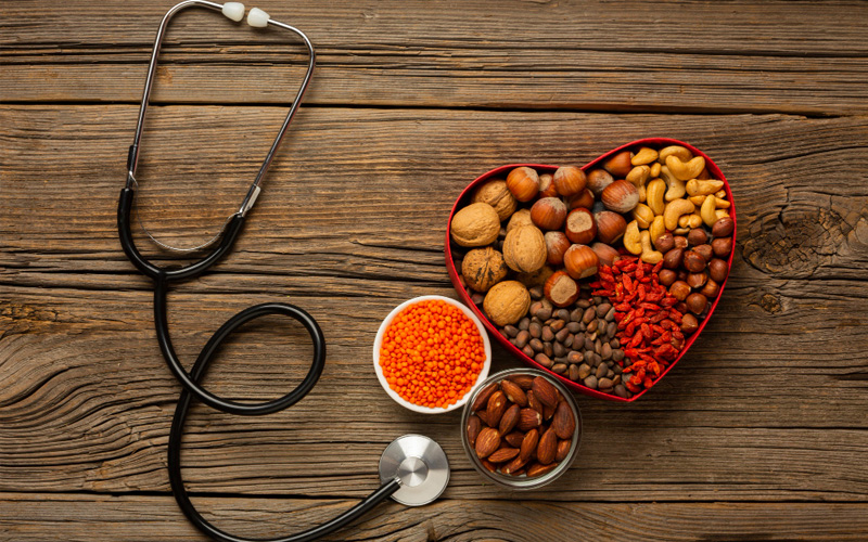 The image shows a collection of various foods and items associated with health and wellness, including nuts, spices, and a red heart-shaped container filled with pills, placed on a wooden surface with a stethoscope and medical cross symbolizing healthcare next to a bowl of carrots.